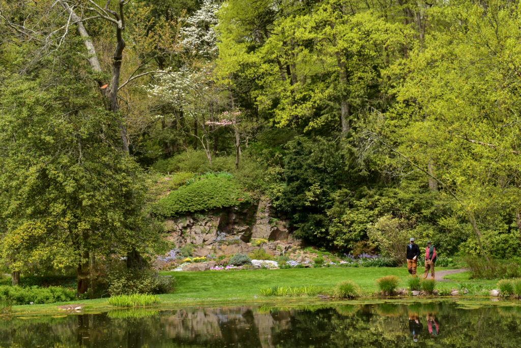 Two people walk a path along a small lake in front of a wooded backdrop with a small rock outcropping.