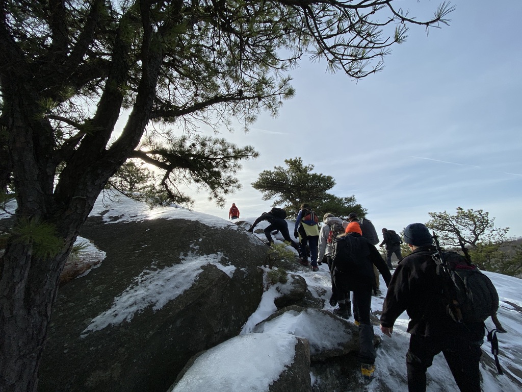 Several warmly dressed hikers climb up a rocky trail with snow on the ground