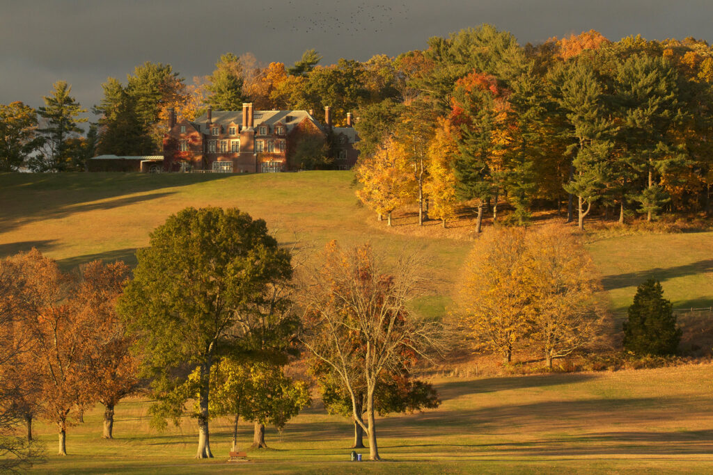 A brick estate sits atop a hill surrounded by trees displaying fall colors.